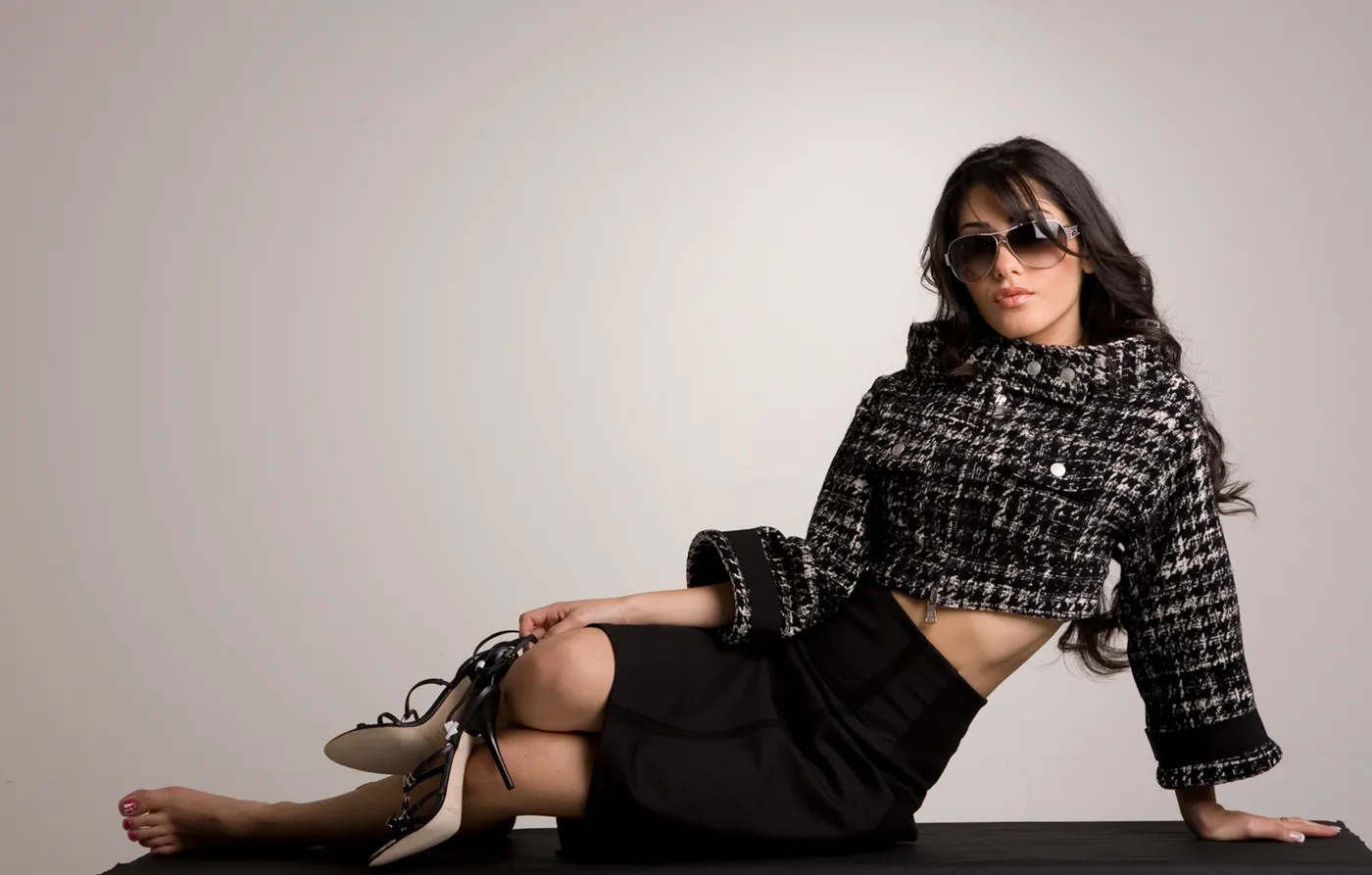 Woman in a cream ribbed off-shoulder knit with oversized dark sunglasses, bare shoulder lit against a grey studio backdrop.